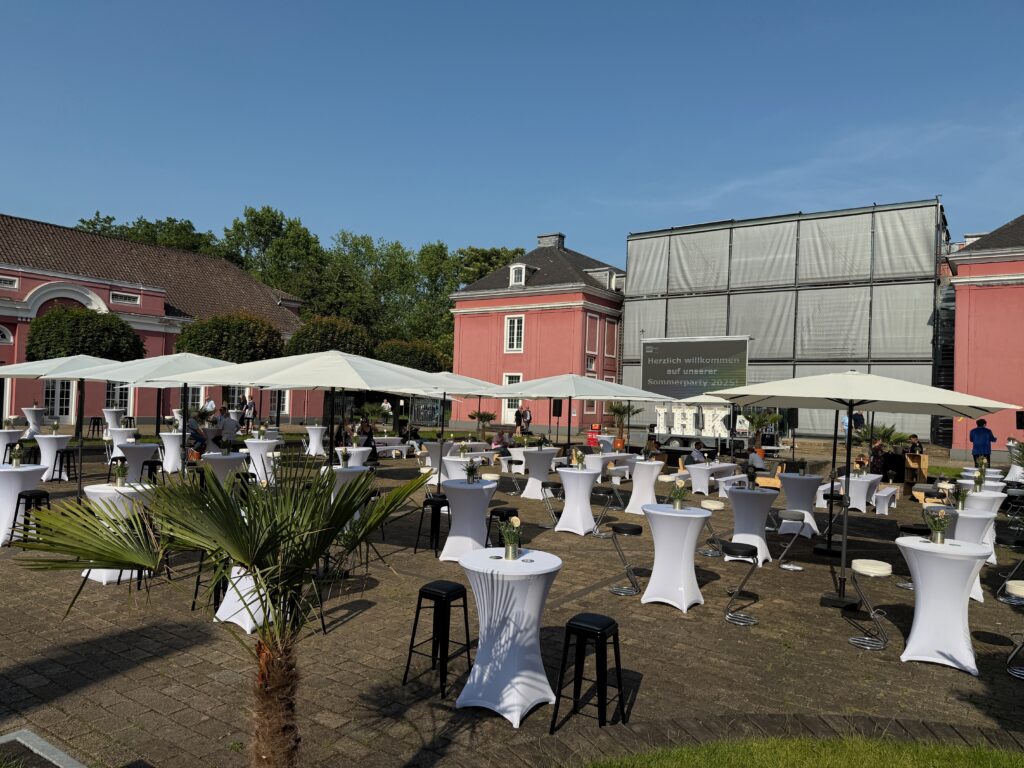 Außenbereich mit Stehtischen, Sonnenschirmen und Palmen vor einem Schloss